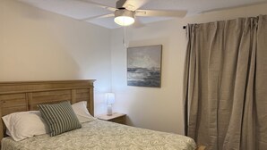 A bedroom with a wooden bed, a nightstand with a lamp, a ceiling fan, a painting, and a window with curtains.