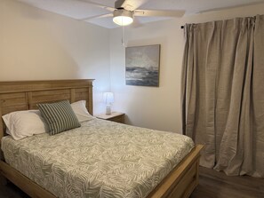 A bedroom with a wooden bed, a nightstand with a lamp, a ceiling fan, a painting, and a window with curtains.