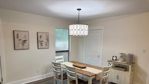 Un comedor con mesa de madera y sillas blancas, una lámpara colgante, y un armario blanco con cafetera y cesta.