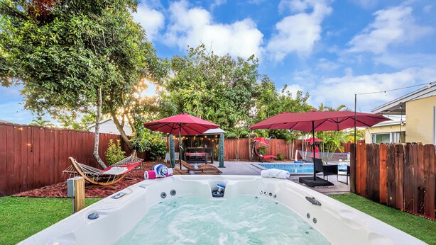Un patio trasero con un jacuzzi, dos sombrillas rojas, una hamaca y una piscina.