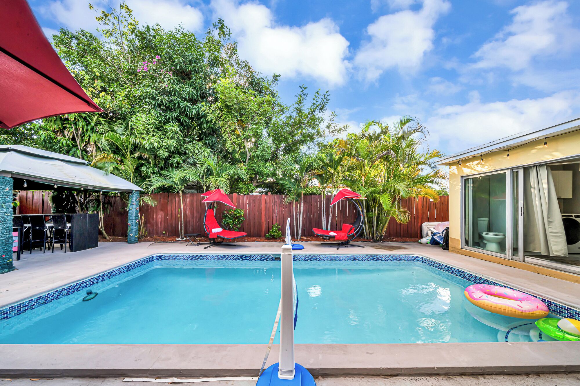 A backyard with a swimming pool, two red lounge chairs, and a covered patio area.
