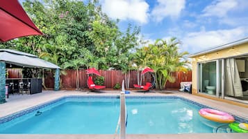 A backyard with a swimming pool, two red lounge chairs, and a covered patio area.