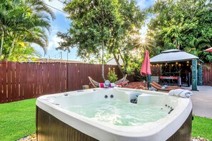 A hot tub in a backyard with a wooden fence, trees, and a patio area with string lights.