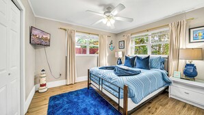 Un dormitorio con cama, ventilador de techo, ventana con cortinas, mesita de noche y lámpara.