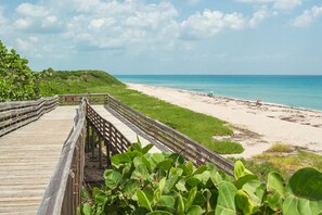 A wooden boardwalk leading to a beach with clear turquoise water and a grassy area.