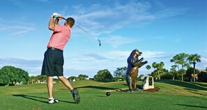 A golf course with a golfer in mid-swing, a bear statue, and a clear blue sky.
