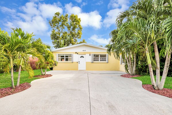 A single-story yellow building with a white door and windows, surrounded by palm trees and a clear sky.