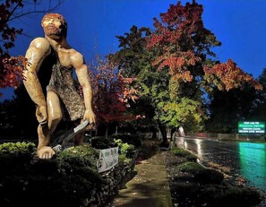 Eine Skulptur einer muskulösen Figur mit einem Schild, auf dem 'Caveman Club' steht, in einer Parkanlage mit Bäumen und einem Weg.