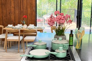 A dining area with a wooden table set for four, a stove with pots and pans, and a view of a balcony with flowers and greenery. - Woodland Hills Hidden Waterfall Oasis | Pool & BBQ (Los Angeles)