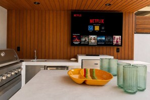 A modern kitchen with a built-in Netflix screen displaying various shows, a grill, and a bowl with utensils. - Woodland Hills Hidden Waterfall Oasis | Pool & BBQ (Los Angeles)