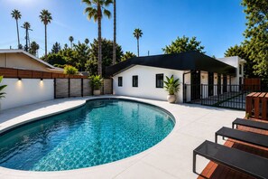 A modern outdoor swimming pool area with a curved design, surrounded by a white building and a wooden deck. - Woodland Hills Hidden Waterfall Oasis | Pool & BBQ (Los Angeles)