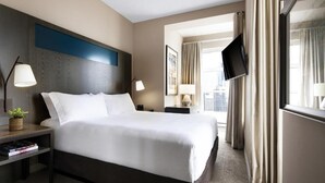 A hotel room with a large bed, two bedside tables with lamps, a flat-screen TV, a window with curtains, and a view of the city. - Dominion Grand Suites Toronto (Toronto)