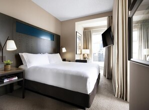 Een hotelkamer met een groot bed, twee nachtkastjes met lampen, een flatscreen televisie, een raam met gordijnen en uitzicht op de stad.