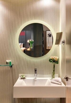 Een badkamer met een ronde spiegel, een witte wastafel, een vaas met bloemen en een handdoekrek.