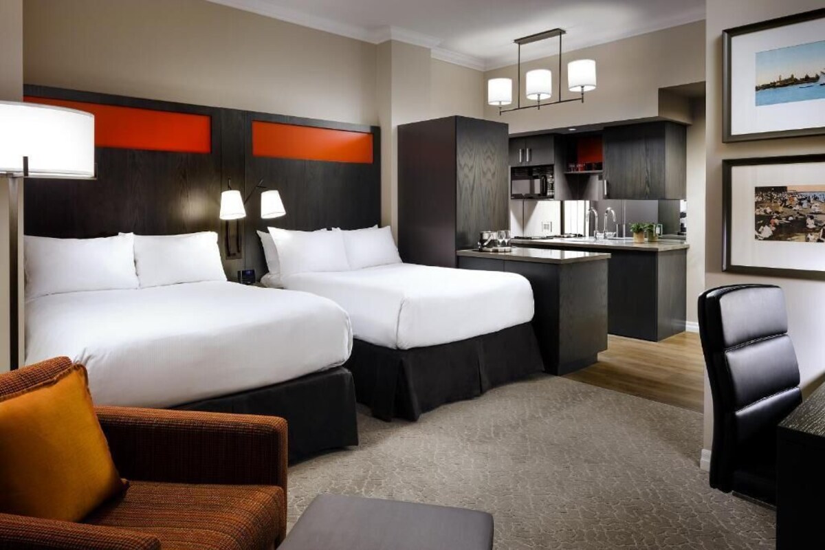 Een hotelkamer met twee bedden, een kitchenette en een zithoek met een fauteuil en poef.