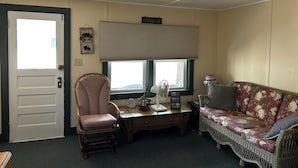 Ein Zimmer mit Tür, Schaukelstuhl, Schreibtisch mit Ventilator, Sofa und Fenster mit Jalousien.