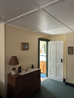A room with a wooden dresser, a lamp, a framed picture, and a door leading to a kitchen.