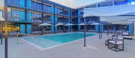 A hotel pool area with a swimming pool, lounge chairs, umbrellas, and a multi-story building with balconies.