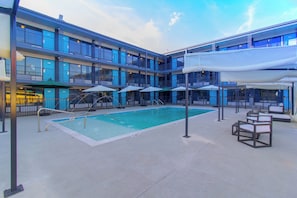 Zona de piscina de hotel con piscina, sillas de descanso, sombrillas y un edificio de varios pisos con balcones.