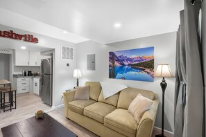 A modern living room with a beige sofa, a coffee table, and a painting of a mountain landscape. - Backyard Breeze: Modern 1BR with Walk-In Shower Near BNA (Nashville)