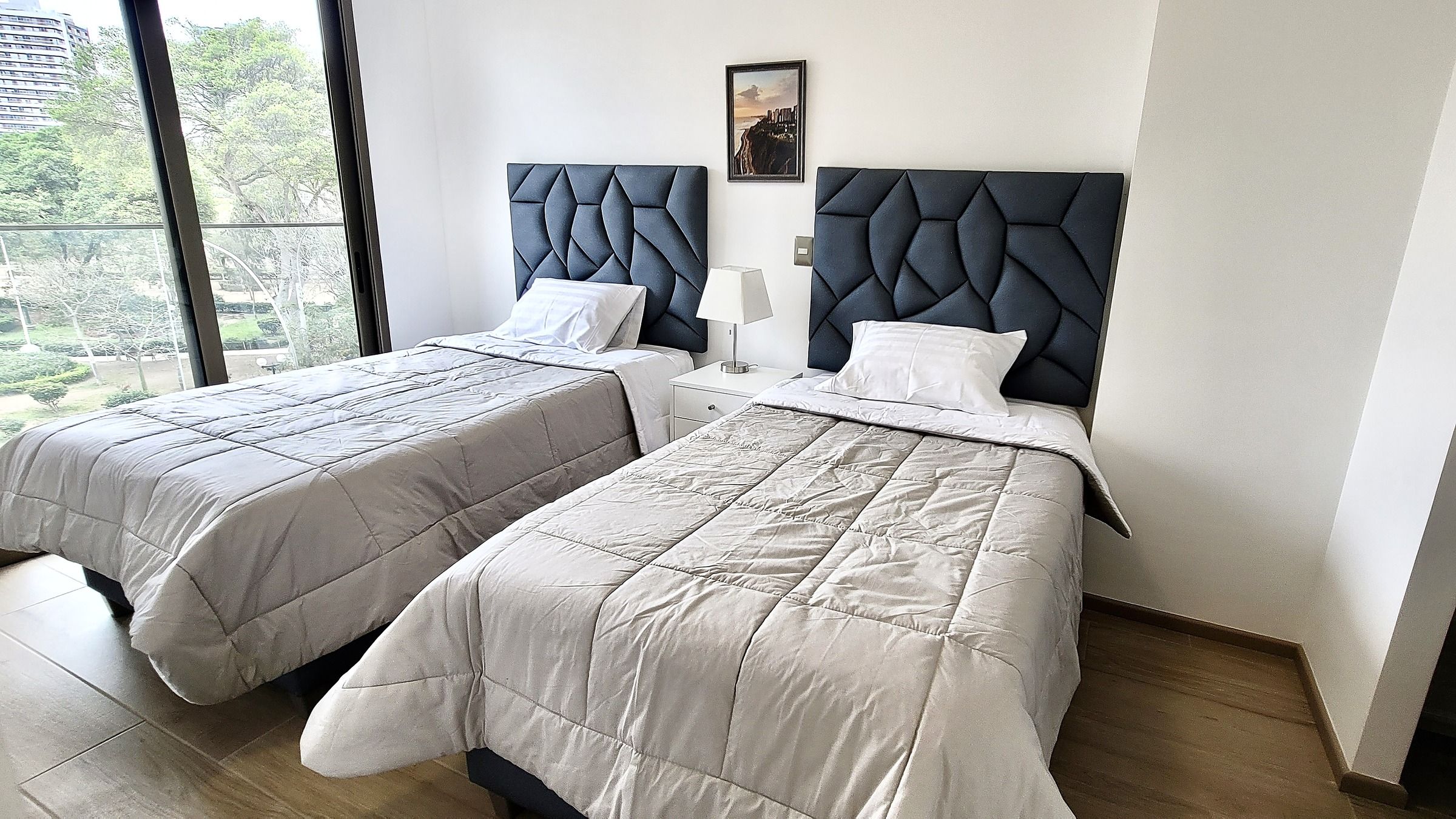 A hotel room with two beds, a view of the outdoors, and a framed picture on the wall.