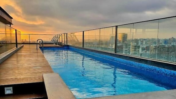 Ein Pool auf dem Dach mit Blick auf die Stadt, umgeben von Glasgeländern und Holzstufen.