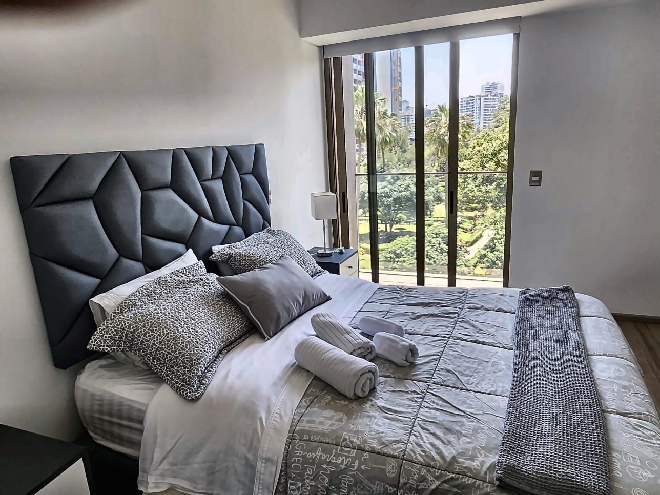 A modern bedroom with a large bed, a headboard, and a view of greenery through a window.