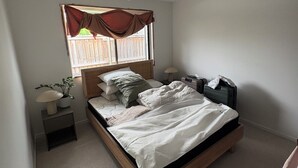 Ein Schlafzimmer mit einem Bett, einem Deckenventilator, einem Fenster mit Vorhängen, einer Nachttischlampe und einem kleinen Tisch mit einer Pflanze.