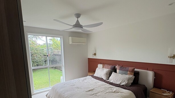Ein Schlafzimmer mit einem großen Bett, einem Deckenventilator, einem Nachttisch und Blick auf eine grüne Außenanlage.