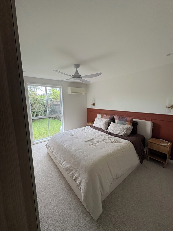 A bedroom with a large bed, a ceiling fan, a nightstand, and a view of a green outdoor area. - Coastal short term rental (Golden Beach)