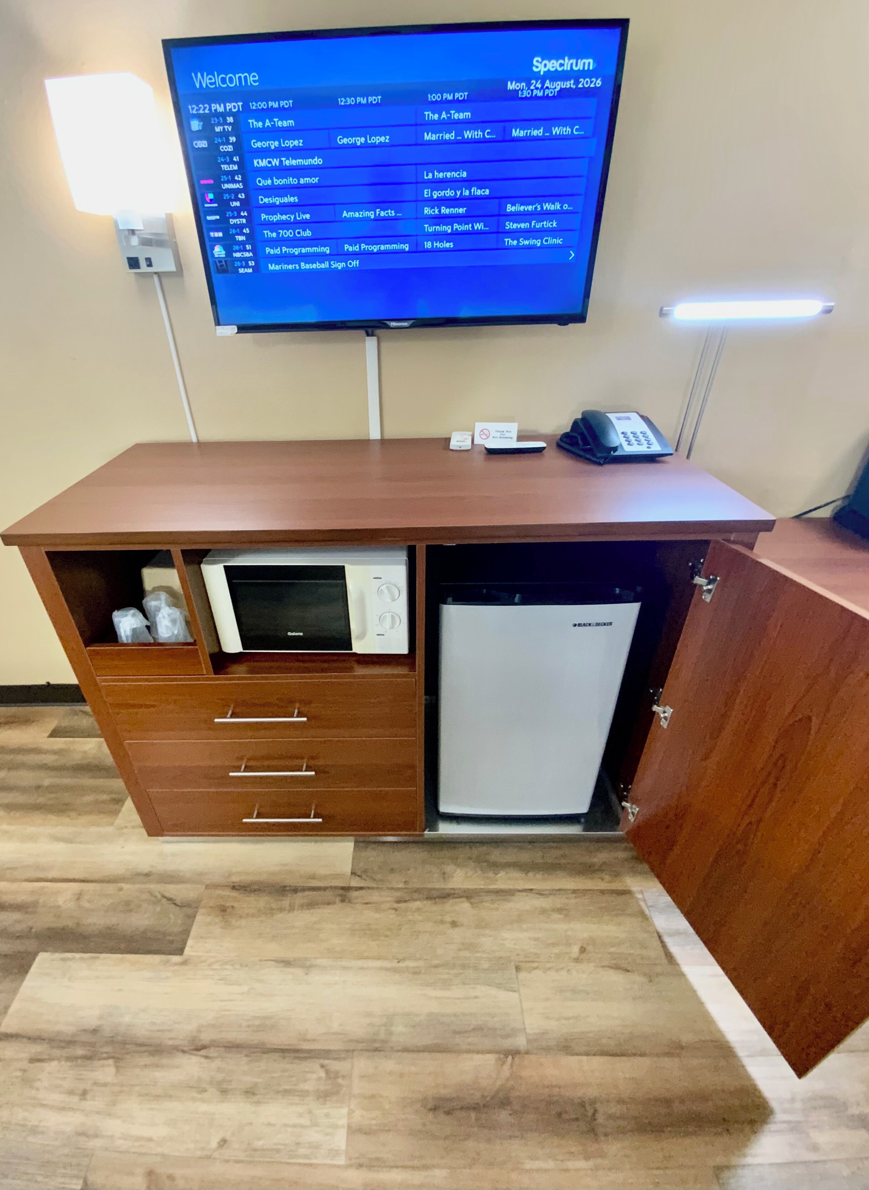 A hotel room with a television displaying a schedule, a desk with a telephone, and a built-in mini-fridge.