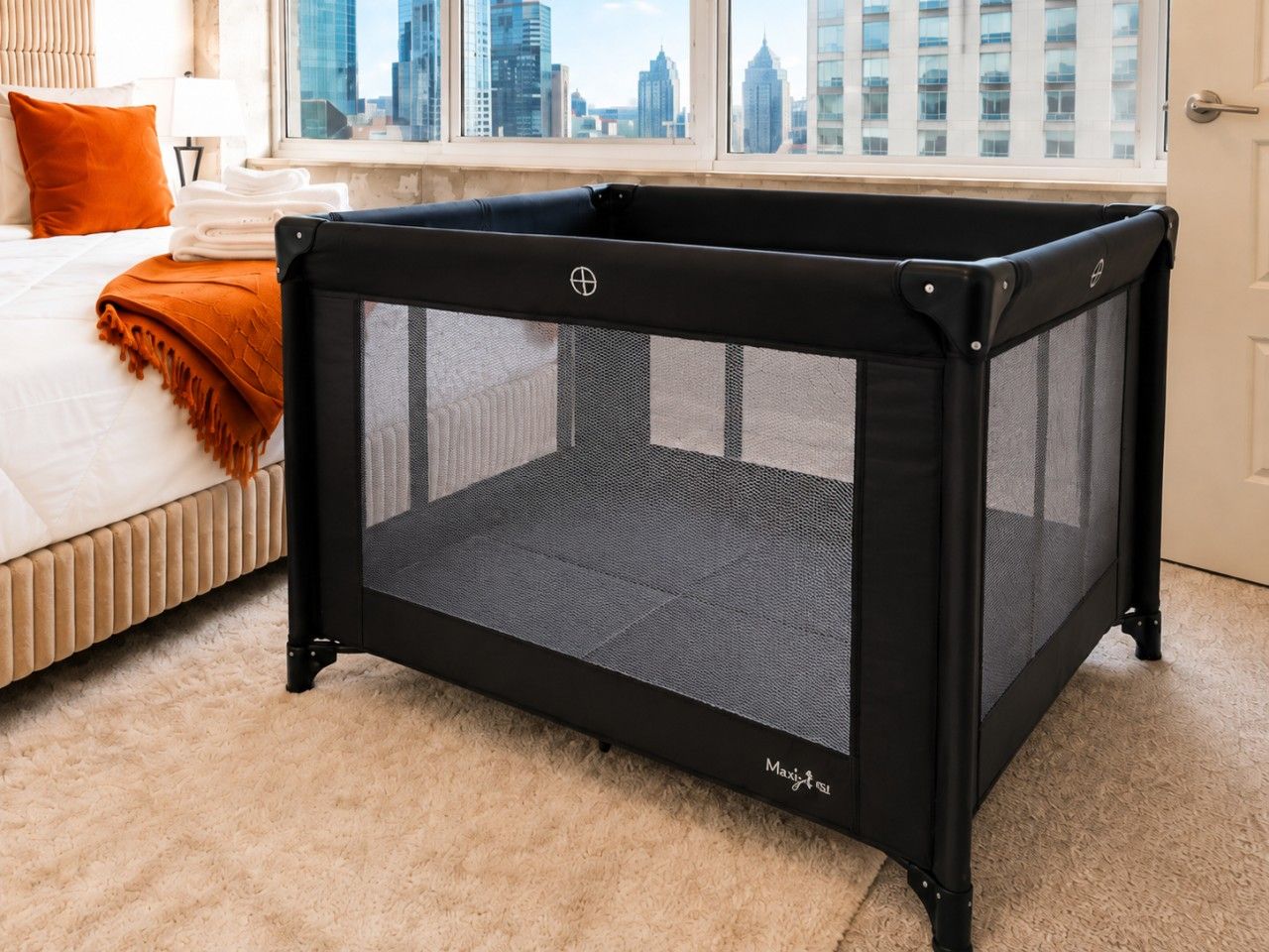 Una valla de juegos negra para bebés con parte superior de malla, ubicada en una habitación que tiene una cama, una ventana con vista a la ciudad y un piso alfombrado.