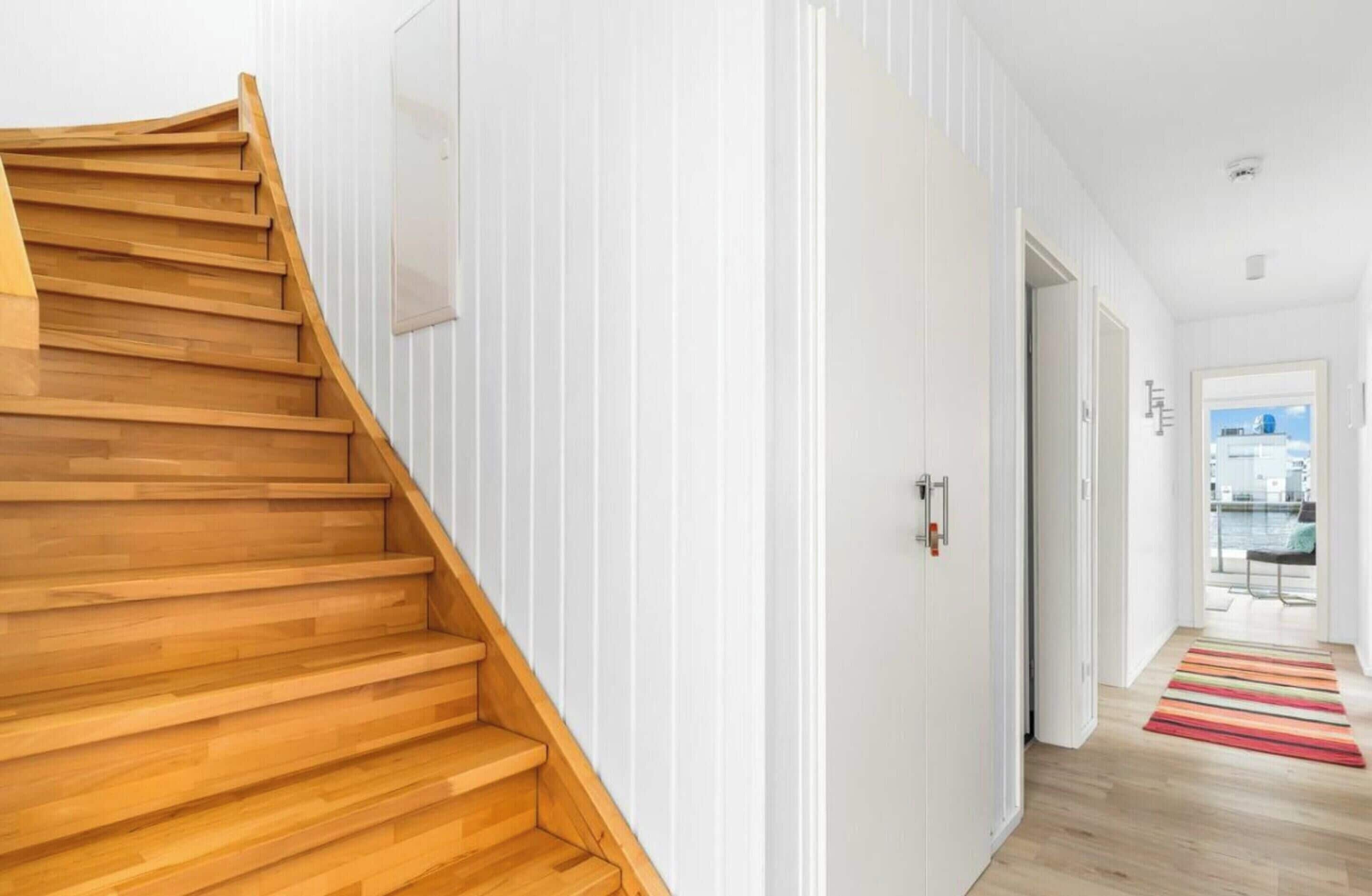 Un intérieur moderne avec un escalier en bois menant à un palier, des murs blancs et une vue sur l’extérieur par une porte ouverte.