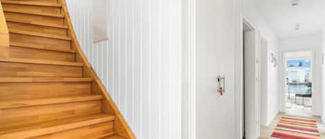 Un intérieur moderne avec un escalier en bois menant à un palier, des murs blancs et une vue sur l’extérieur par une porte ouverte.