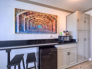 Una cocina con encimera negra, microondas, refrigerador y un cuadro de un túnel en la pared.