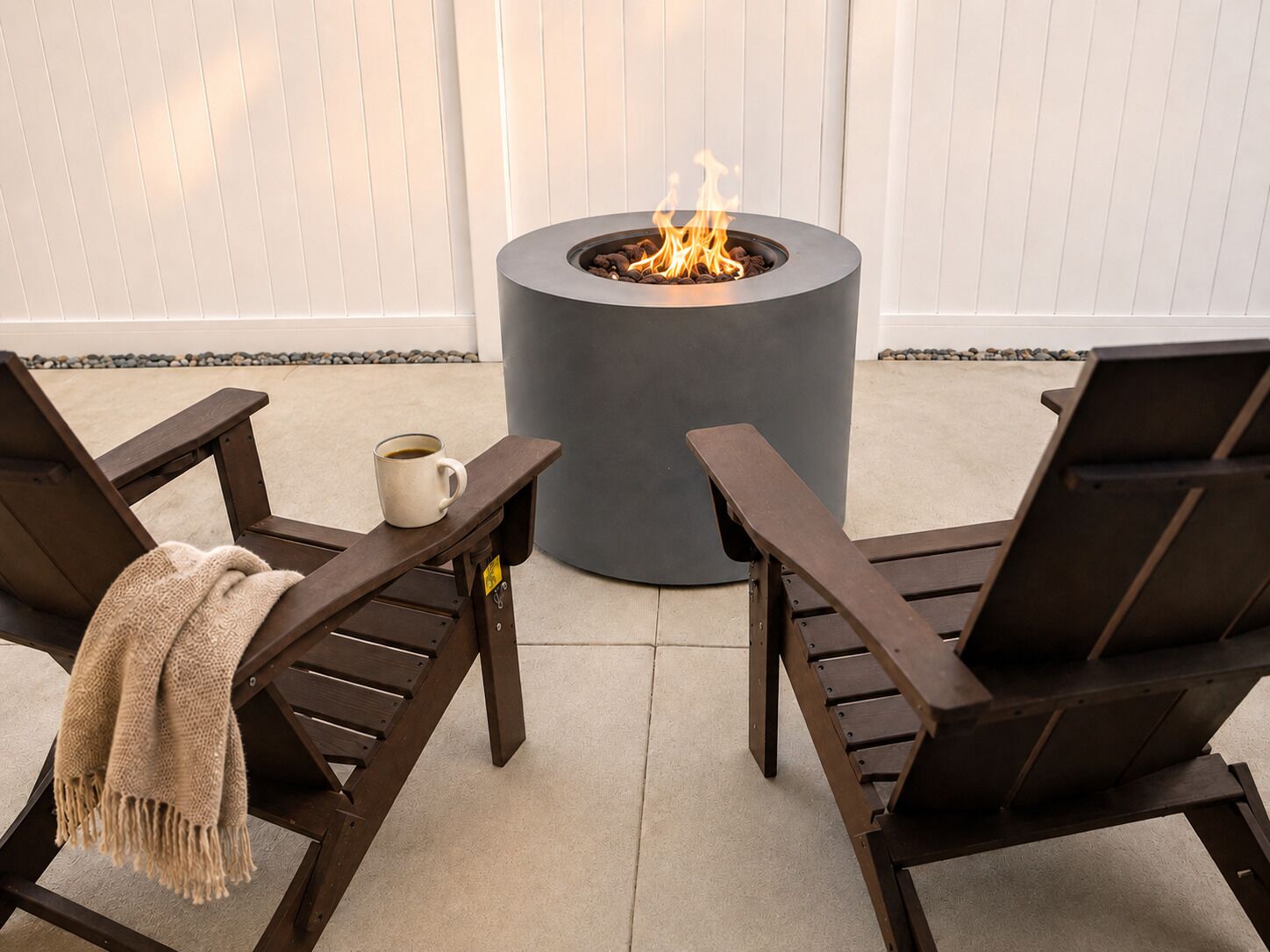Two wooden lounge chairs with a coffee cup and a blanket, a round fire pit with flames, and a white fence in the background.