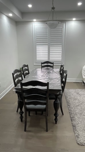 Ein Esszimmer mit einem großen Tisch, umgeben von Stühlen, einer Chandelüster und einem Fenster mit Jalousien.