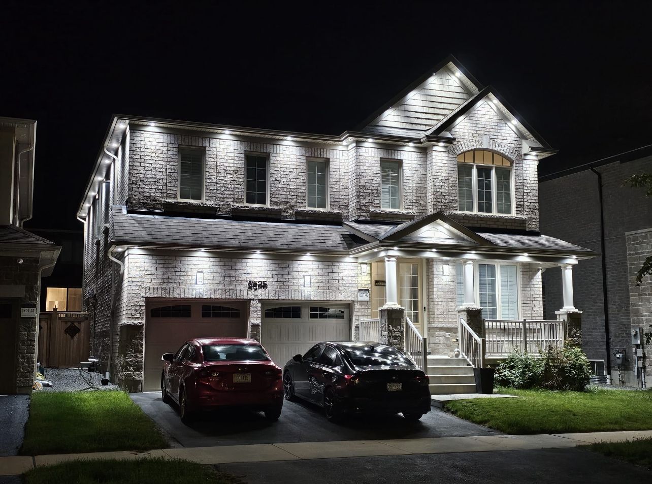 A two-story house with a garage, two cars parked in front, and a well-lit exterior.