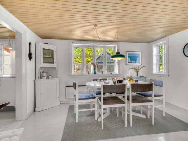 A dining area with a white table, chairs, and a rug. There are windows with views of greenery outside.