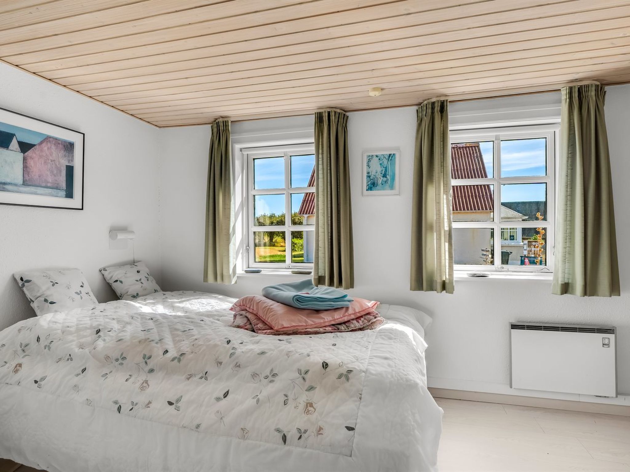 Un dormitorio con una cama, dos ventanas, un radiador y un cuadro en la pared.