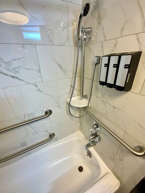 Un baño con bañera, barras de apoyo, cabezal de ducha manual y dispensador de jabón fijado a la pared.