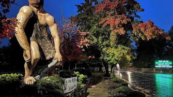 A sculpture of a muscular figure with a sign reading 'Caveman Club' in a park setting with trees and a pathway.