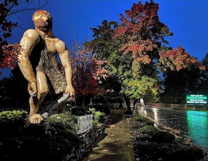 En skulptur af en stærk figur med et skilt, der siger 'Caveman Club', i en park med træer og en sti.