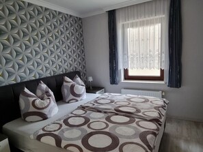 A modern bedroom with a geometric patterned wall, a bed with circular patterned bedding, a nightstand, and a window with sheer curtains. - Franzi Holiday Apartment (Heringsdorf)