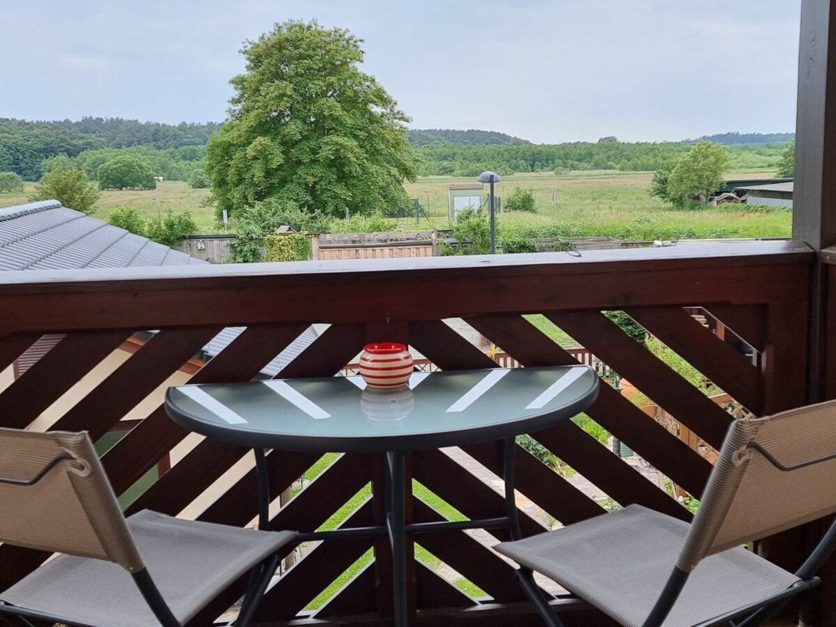 A balcony with a table and two chairs, overlooking a green landscape with trees and a hill.