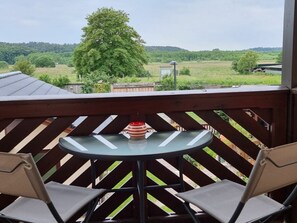 A balcony with a table and two chairs, overlooking a green landscape with trees and a hill. - Franzi Holiday Apartment (Heringsdorf)
