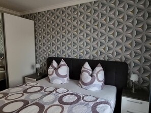 A bedroom with a patterned wall, a bed with a patterned bedspread, two pillows, a nightstand with a lamp, and a drawer unit. - Franzi Holiday Apartment (Heringsdorf)