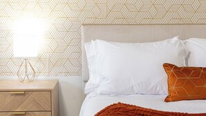 A bedroom with a bed, white bedding, a brown throw blanket, a nightstand with drawers, a lamp, and a geometric patterned wall.