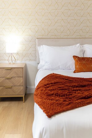A bedroom with a bed, white bedding, a brown throw blanket, a nightstand with drawers, a lamp, and a geometric patterned wall.