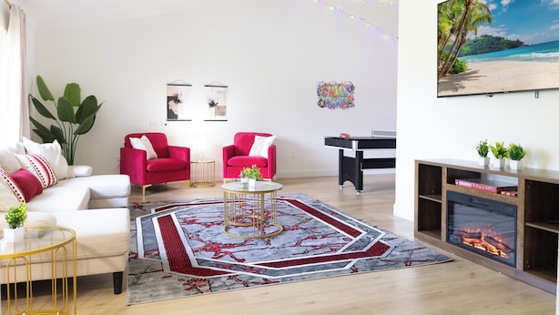 A living room with a ceiling fan, a fireplace, a large painting of a beach, and a red armchair.